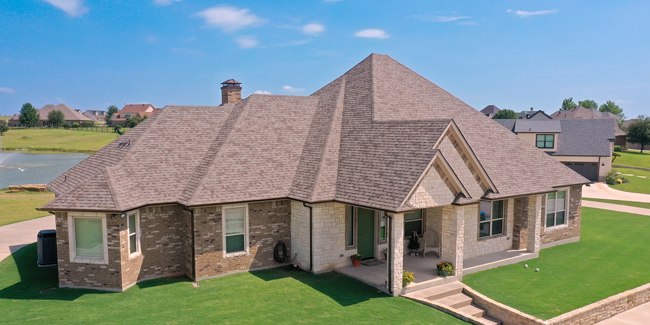 Drone view of a brick and stone home with a tan shingle hip roof, a pond and neighboring homes behind it