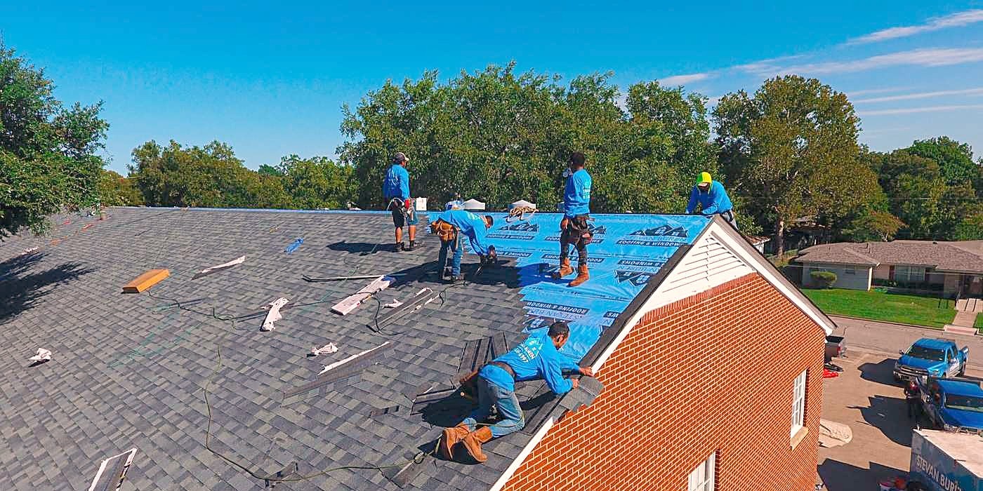 A crew laying blue synthetic underlayment across a stripped roof deck
